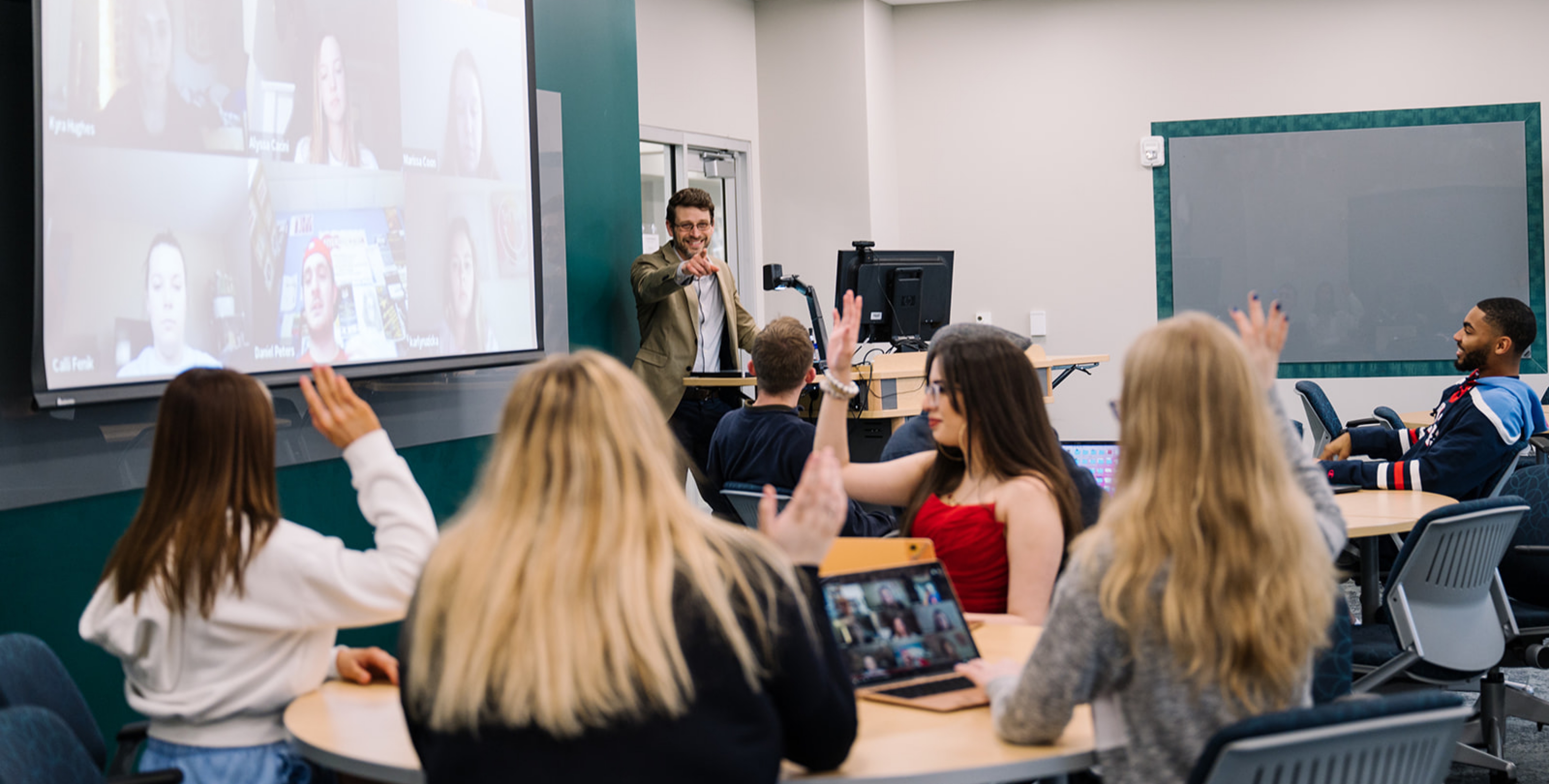 A smiling instructor calls on one of multiple students, whose hands are raised, while remote students look on via a Zoom call.